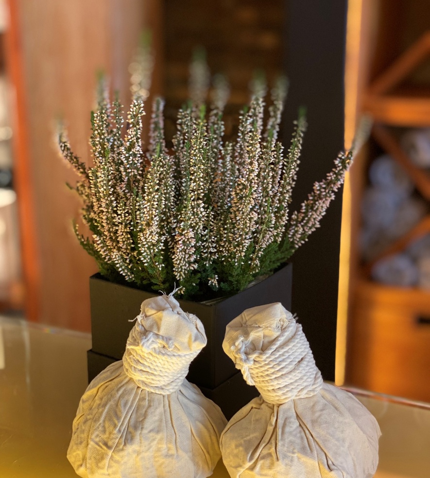 Two cotton herbal massage pouches beside blooming heather flowers in a serene hotel spa setting.