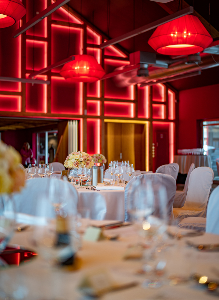 Elegant banquet hall with round tables, white chair covers, floral centerpieces and glassware under warm red geometric wall lighting