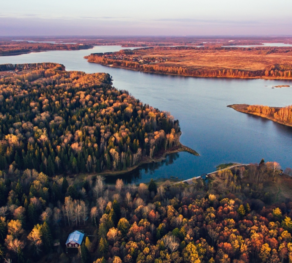 Expansive lake surrounded by golden autumn forests with a small cabin near the shore under soft evening light.