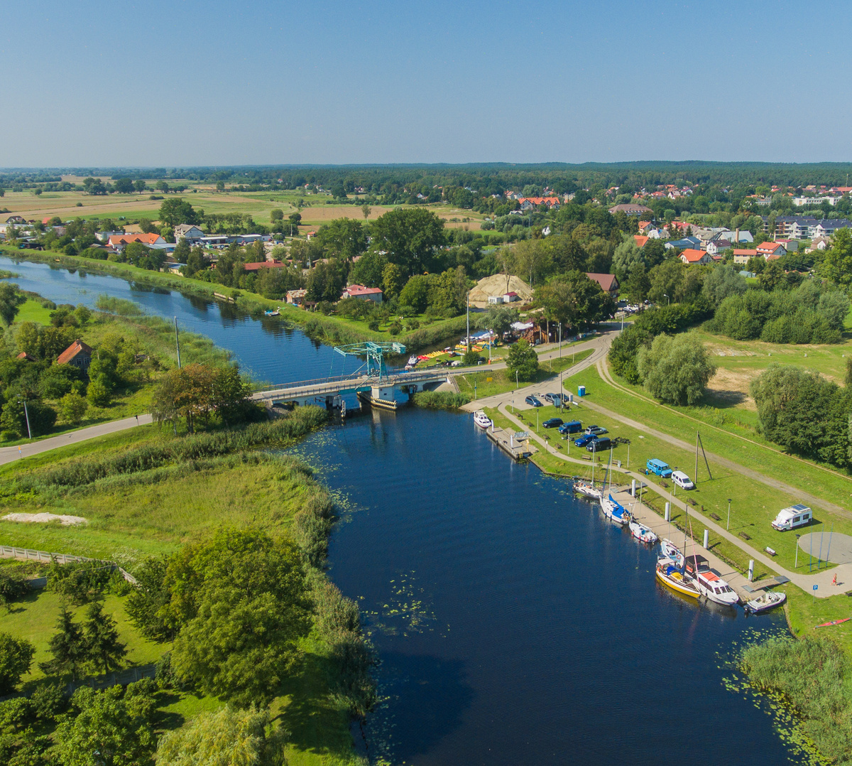 Marina nad rzeką z cumującymi łodziami, małym mostem, parkingiem nad wodą i zielonymi ścieżkami