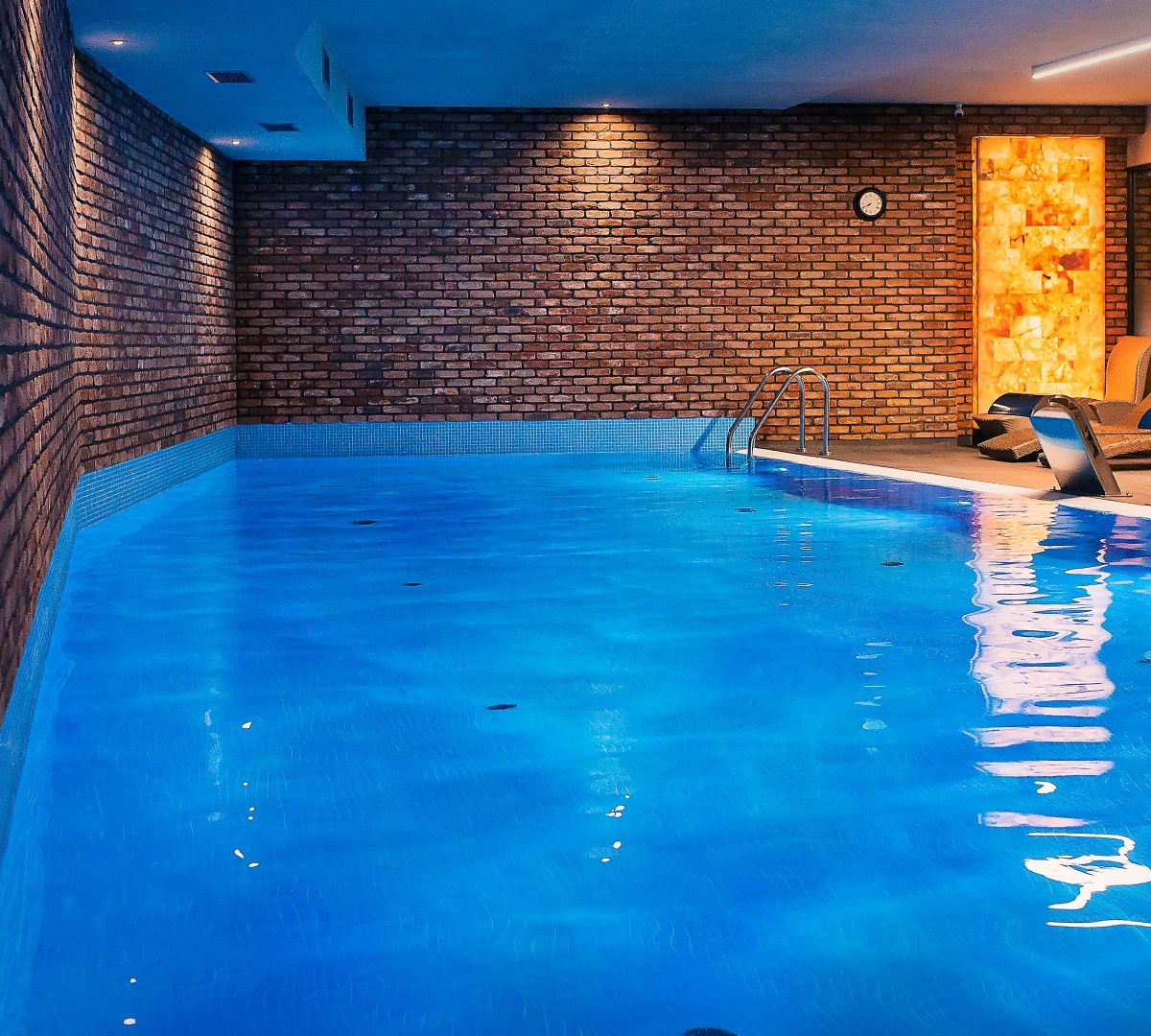 Indoor heated hotel pool with glowing blue water, exposed brick walls, ladder entry, backlit stone panels and poolside loungers