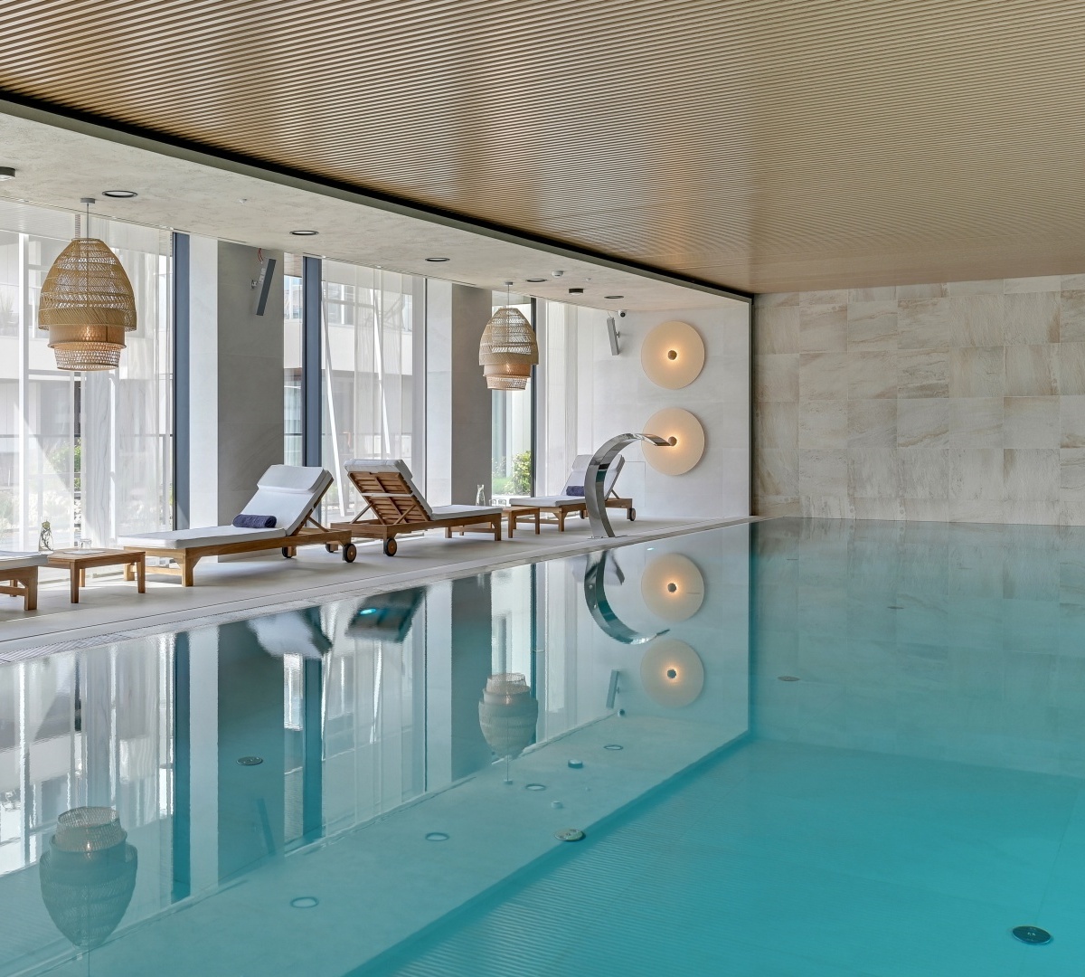 Tranquil indoor pool with turquoise water, wooden loungers, wicker lights, floor-to-ceiling glass and curved water spout