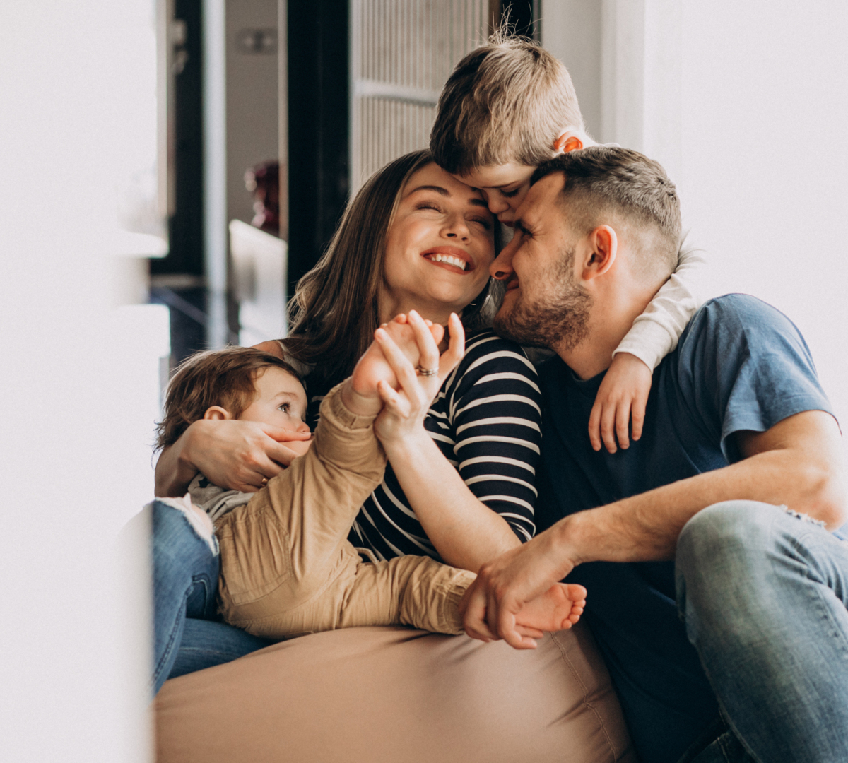 Family relaxing in a bright modern suite living area: parents and two children cuddling on a comfy sofa in warm natural light.