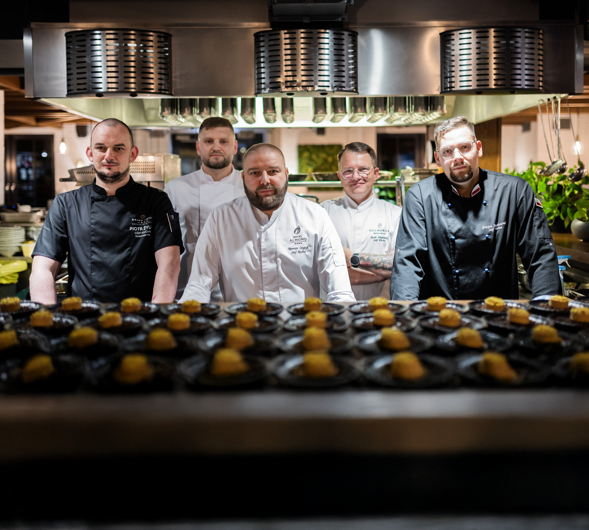Fem uniformerade kockar vid öppet kök, rostfri huva ovan, rader med upplagda smårätter i förgrunden.