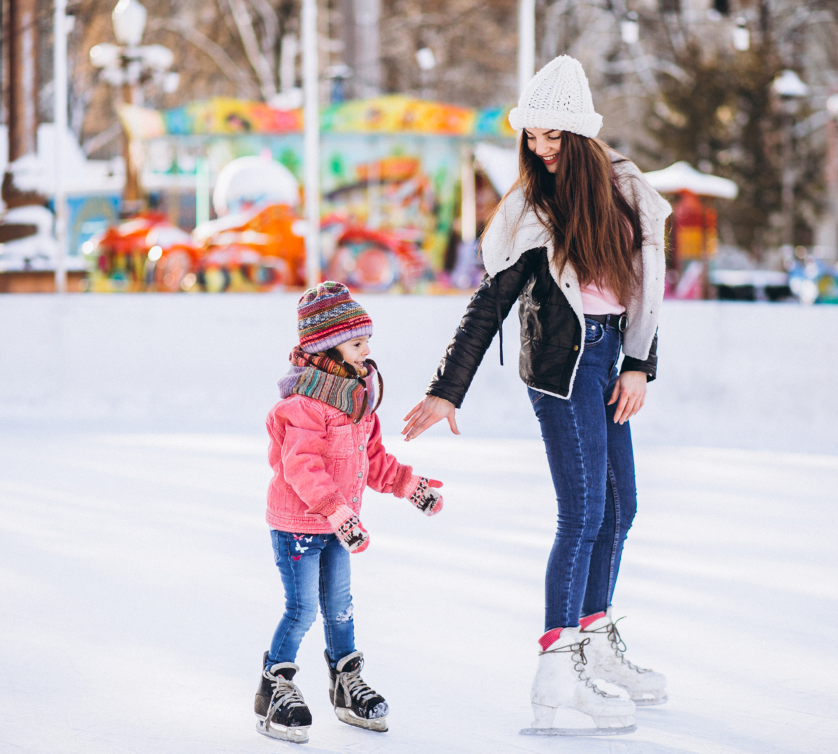 Kobieta pomaga dziecku na zewnętrznym lodowisku, w tle kolorowe karuzele w zaśnieżonym parku