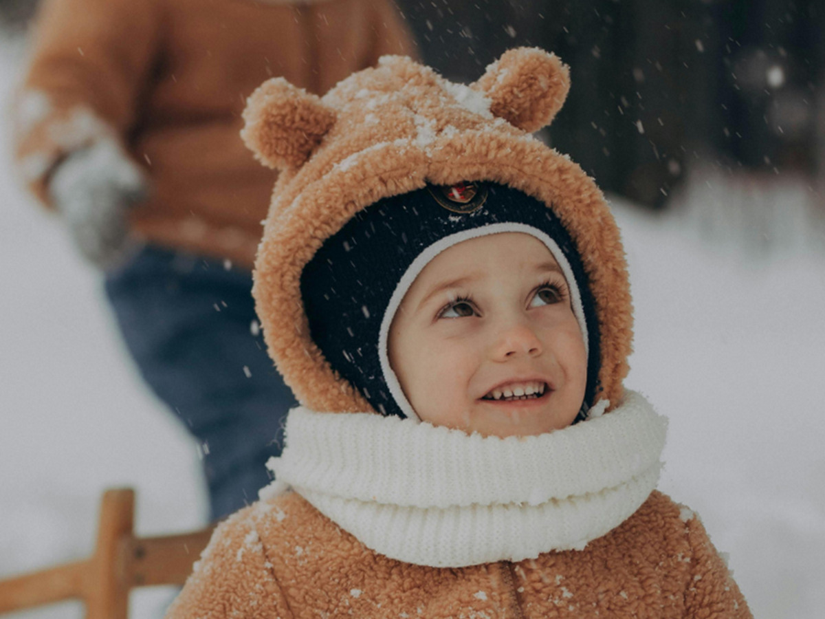Barn i kosehette med ører og hvit strikket skjerf smiler på en treslede i fallende snø