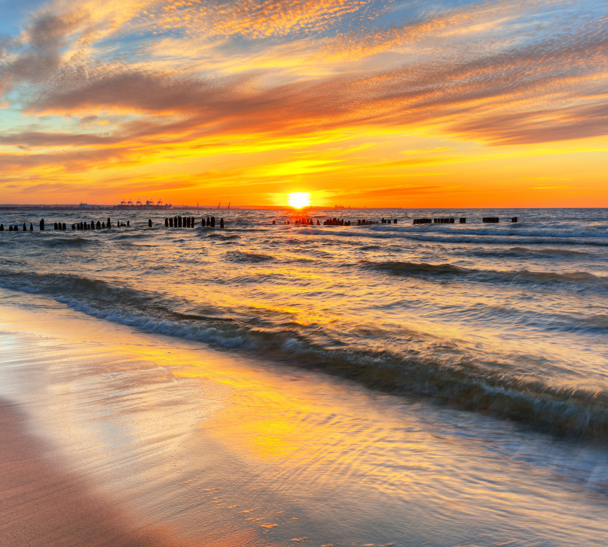 Zachód słońca nad piaszczystą plażą, łagodne fale i drewniane falochrony, złote niebo odbija się na mokrym piasku