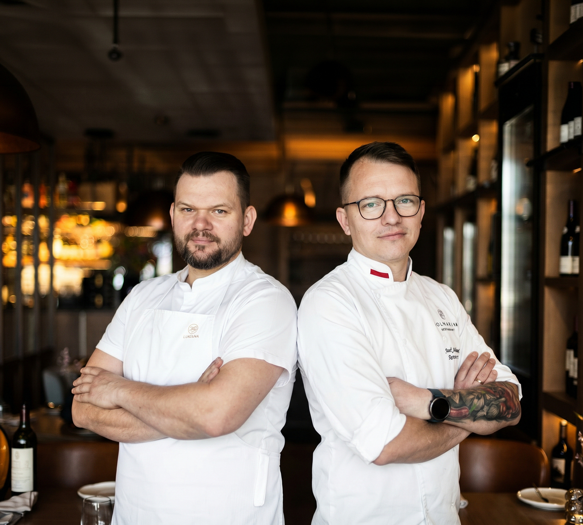 To kokker i hvite uniformer står med armene i kors i en varmt opplyst restaurant ved hyller med vinflasker og intime bord.
