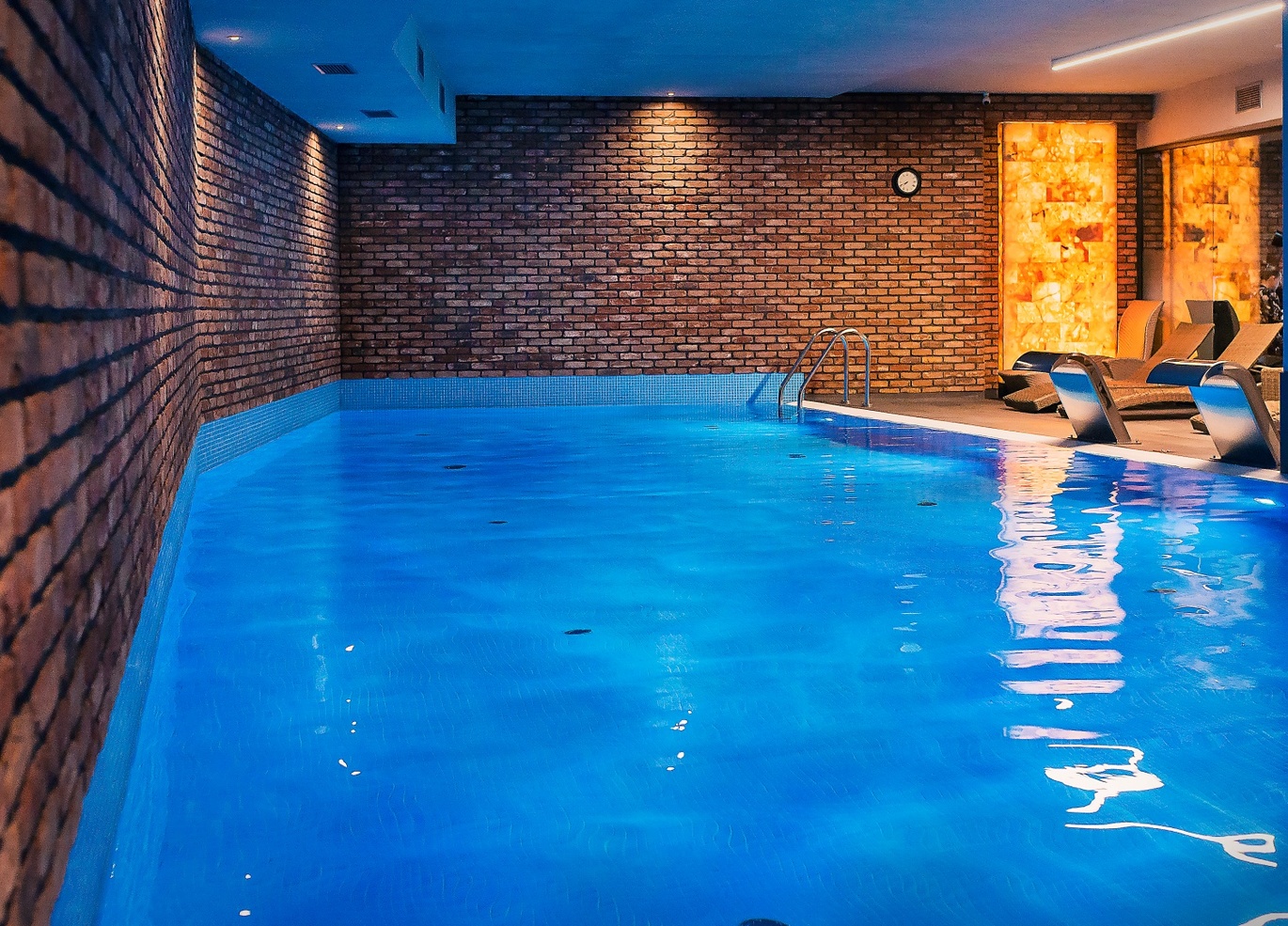Kryty, ogrzewany basen hotelowy z niebiesko podświetloną wodą, ścianami z cegły, drabinką, podświetlanymi panelami kamiennymi i leżakami