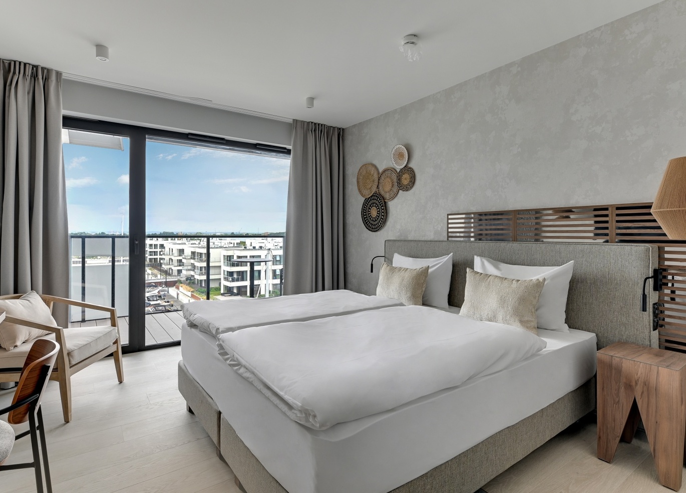 Nowoczesny pokój z dwiema złączonymi łóżkami, białą pościelą, drewnianym zagłówkiem, wiszącymi lampami, biurkiem, fotelem i balkonem z widokiem na nowoczesne budynki nad wodą
