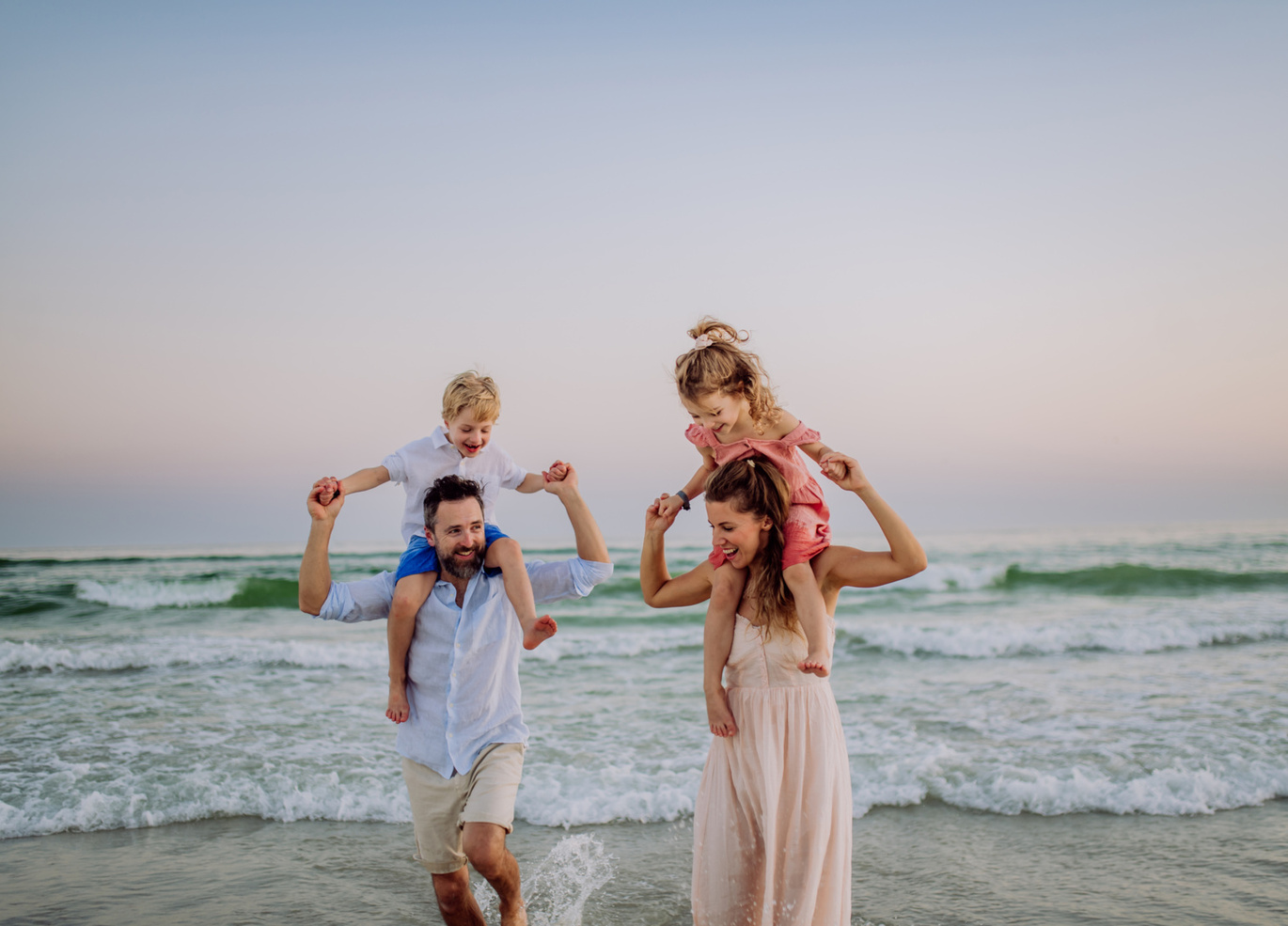 Rodzice niosą dzieci na ramionach, idąc w płytkiej wodzie przy spokojnej plaży o zachodzie słońca.