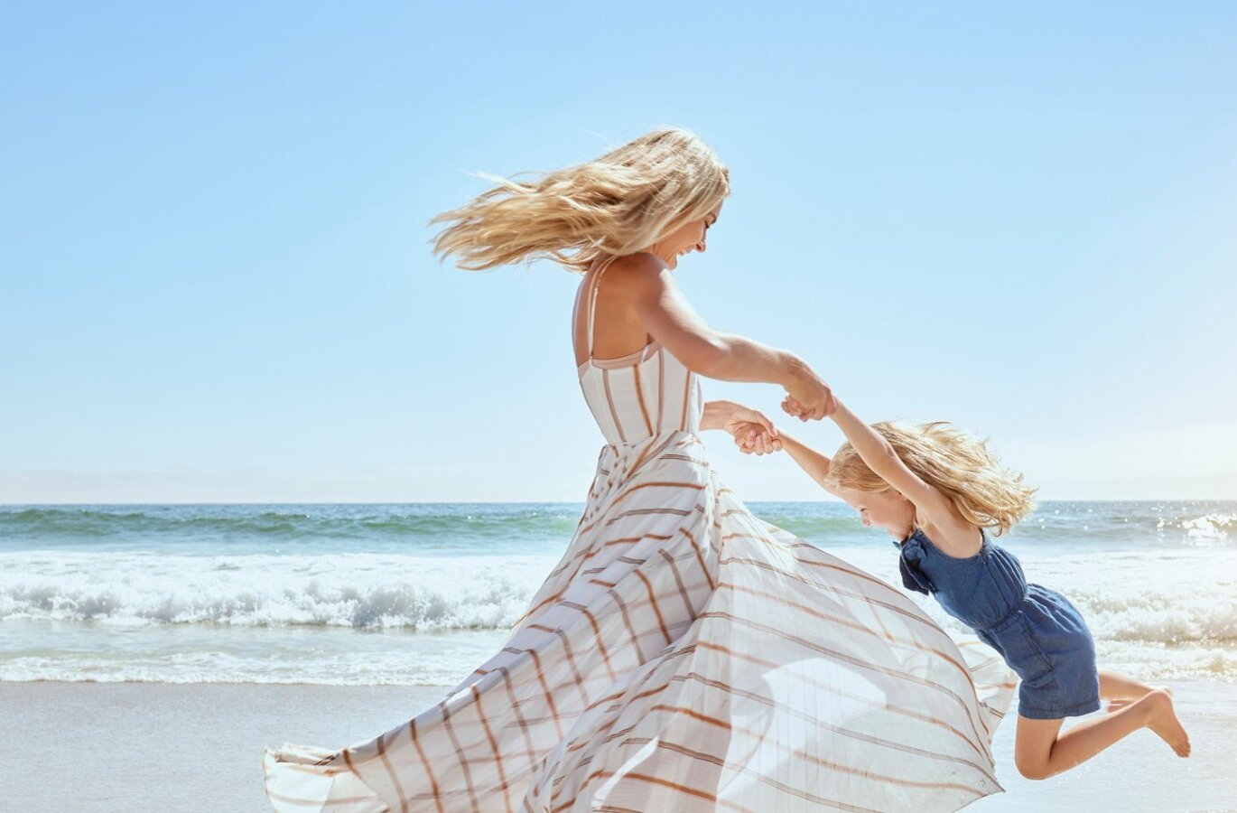 Matka kręci córkę na słonecznej piaszczystej plaży z łagodnymi falami — przyjazny rodzinom kurort