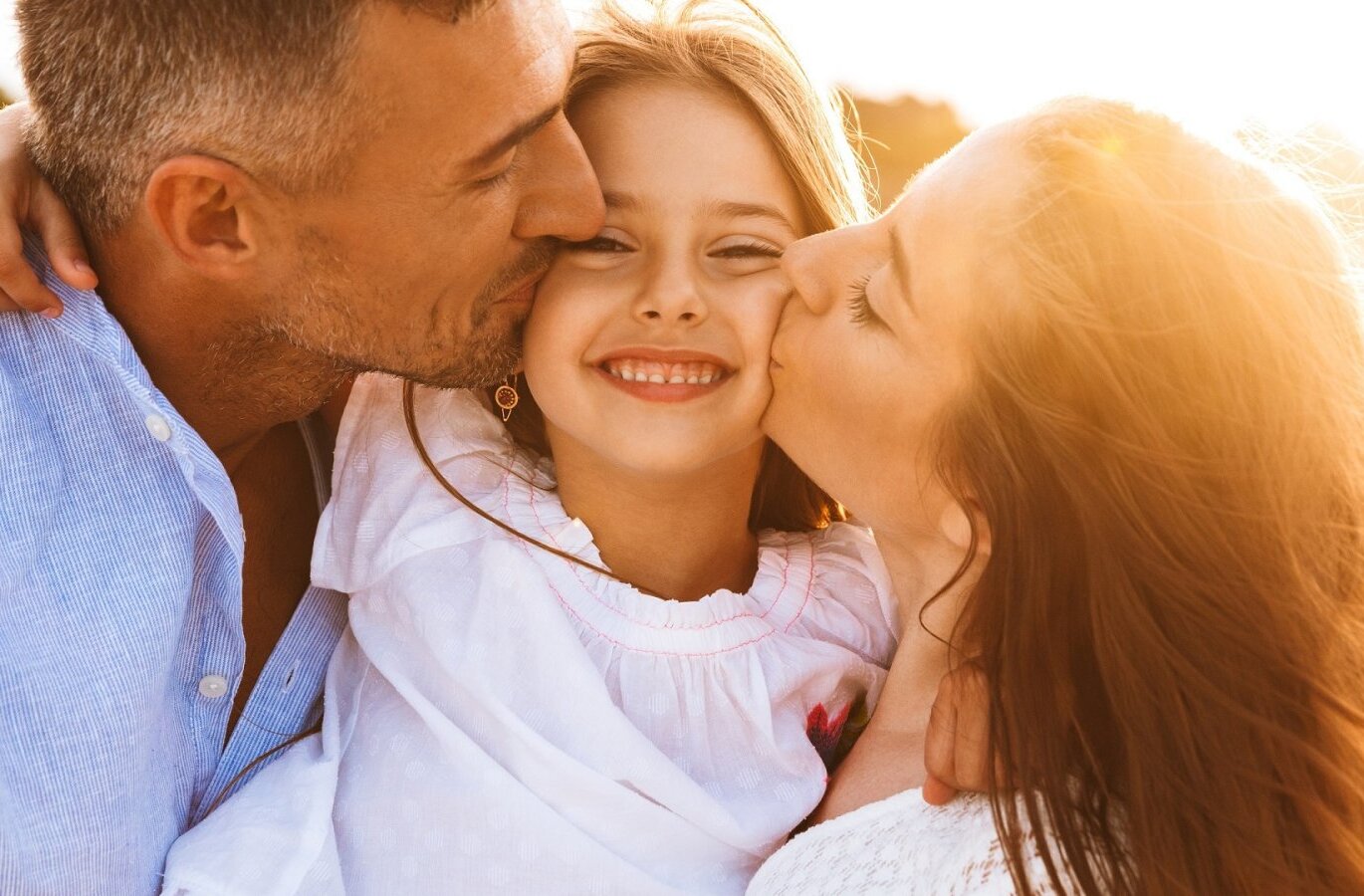 Rodzice całują uśmiechniętą córkę o złotej godzinie na plaży, ciepłe światło i swobodna letnia atmosfera