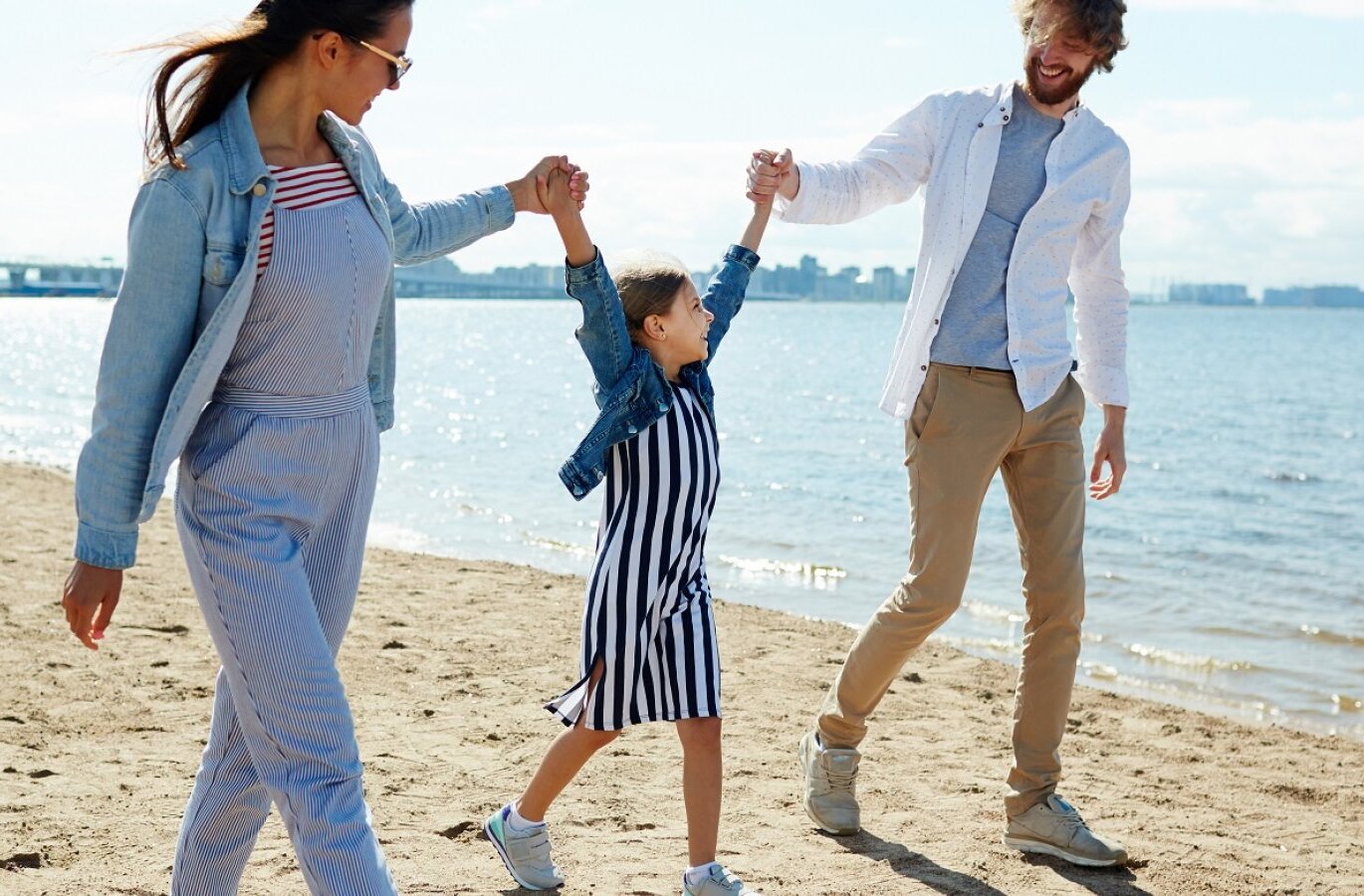 Family walking hand-in-hand along a sunny hotel beachfront with sandy shore, calm sea and distant city skyline