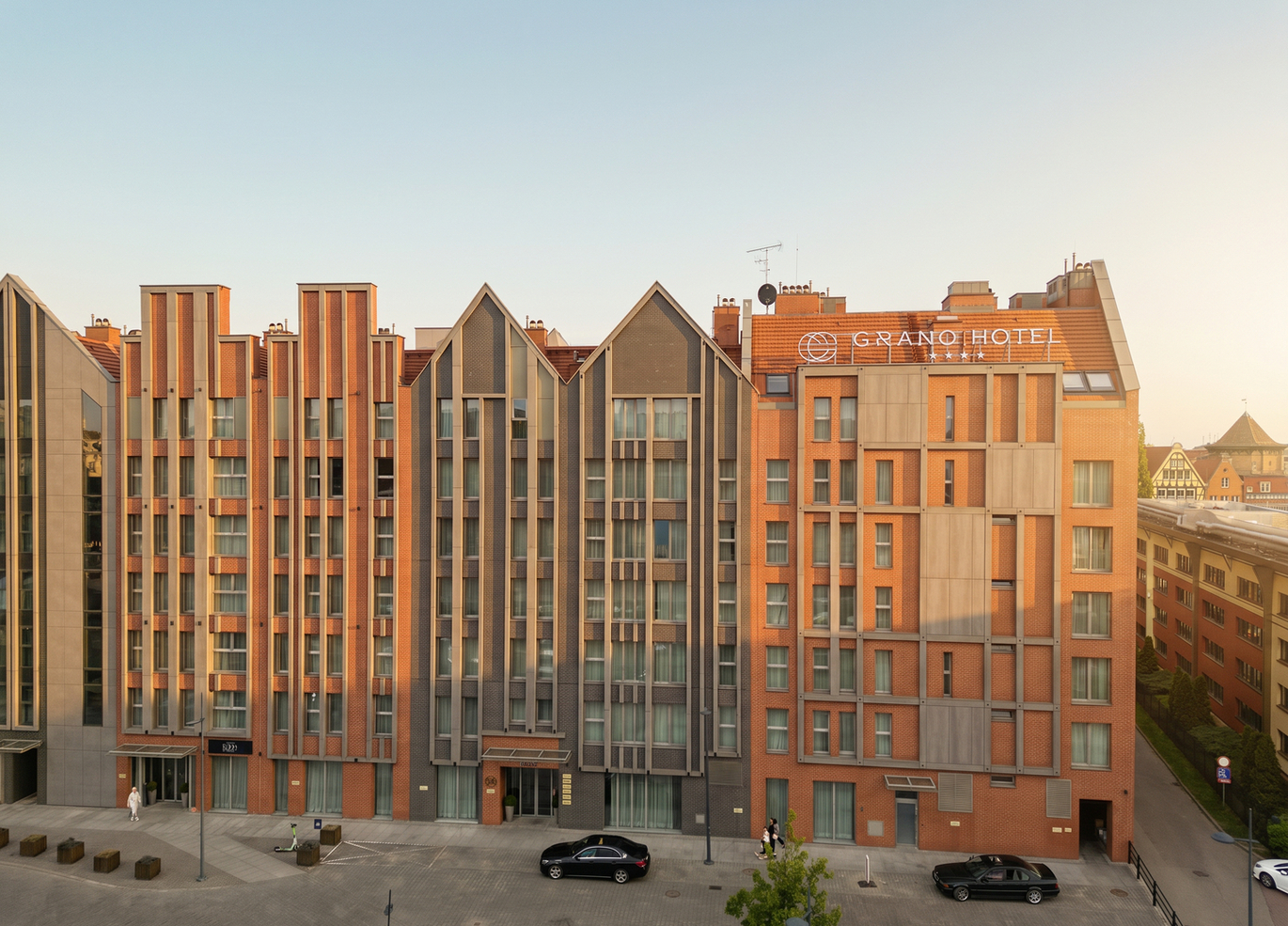 Ceglasty, nowoczesny hotel z ostrym szczytowym dachem, licznymi oknami i wejściem od ulicy w ciepłym świetle zachodu