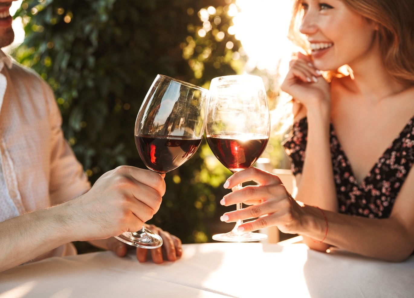 Paar stößt mit Rotwein auf sonnenbeschienener Hotelterrasse an, lächelnde Frau, entspanntes Essen im Freien vor grünem Hintergrund