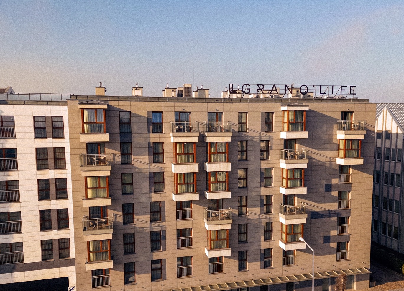 Nowoczesna fasada hotelu z prywatnymi balkonami, dużymi oknami i widocznym szyldem na dachu nad spokojną ulicą