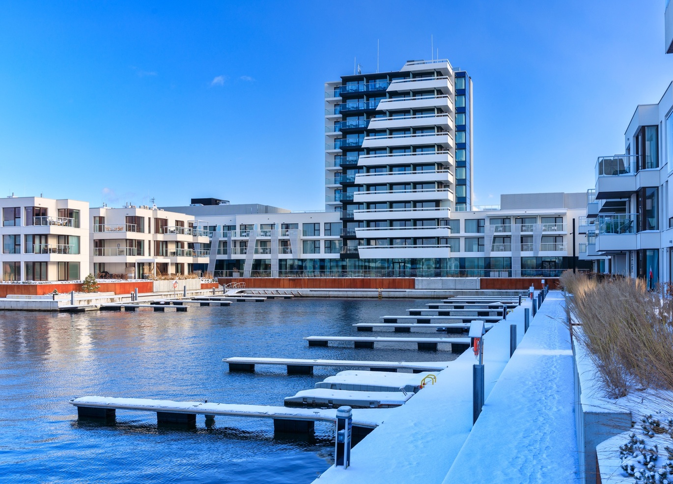 Nowoczesny hotel nad wodą z balkonami i prywatnymi tarasami, mariną i pomostami pokrytymi śniegiem pod błękitnym niebem