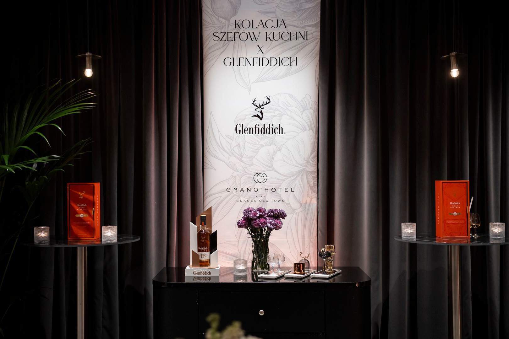 Stoisko degustacyjne Glenfiddich z butelką, kieliszkami, świecami i fioletowymi kwiatami przed banerem Grano Hotel
