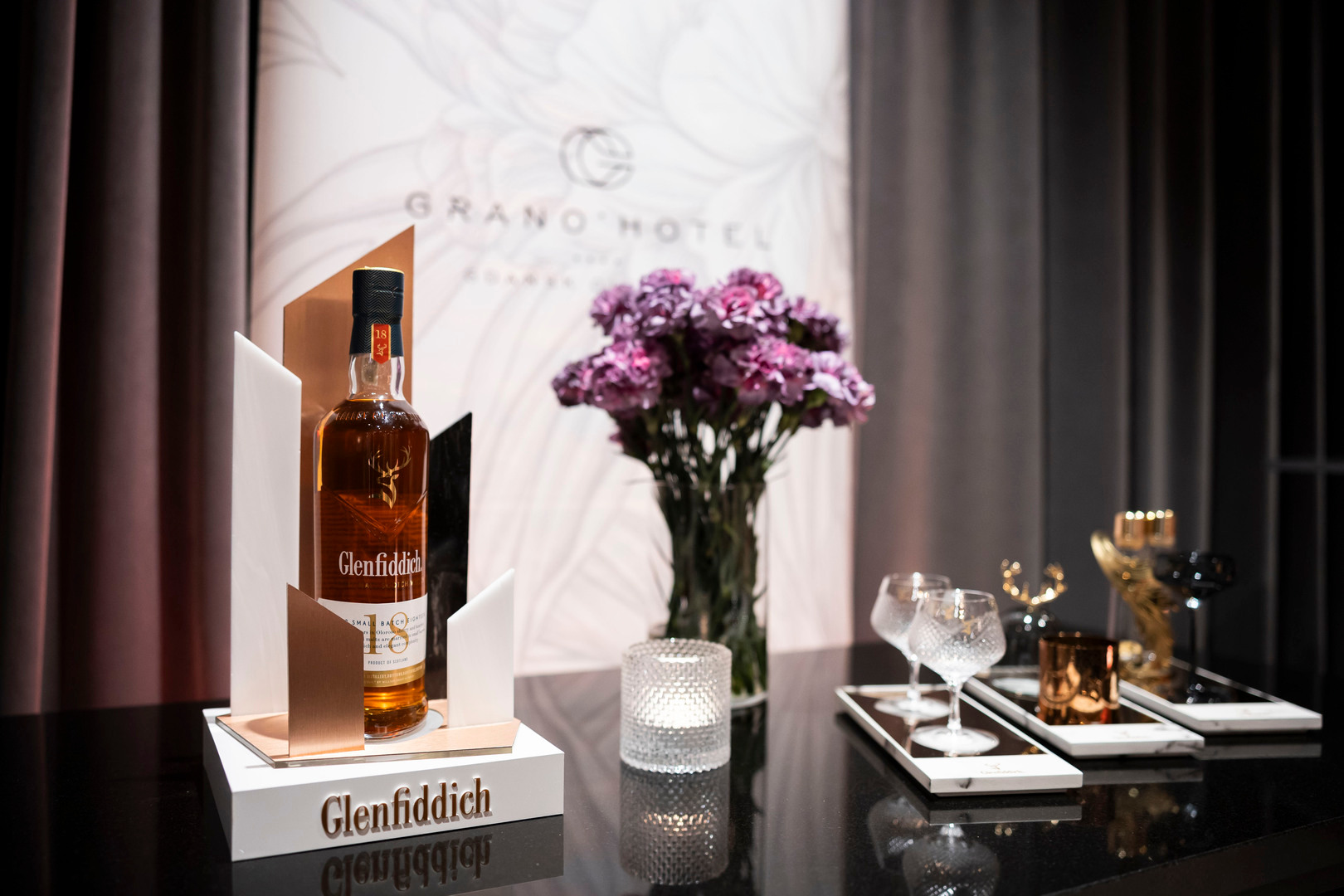 Butelka whisky Glenfiddich na hotelowym barze, kryształowe kieliszki, świeca i fioletowe goździki, elegancki salon