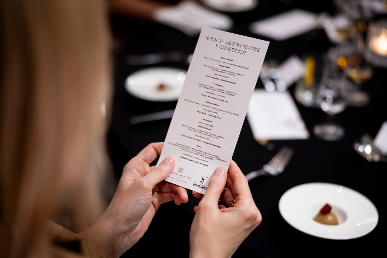 Dłonie trzymają kartę degustacyjną podczas eleganckiej kolacji przy świecach; czarny obrus, przystawka i kieliszki.