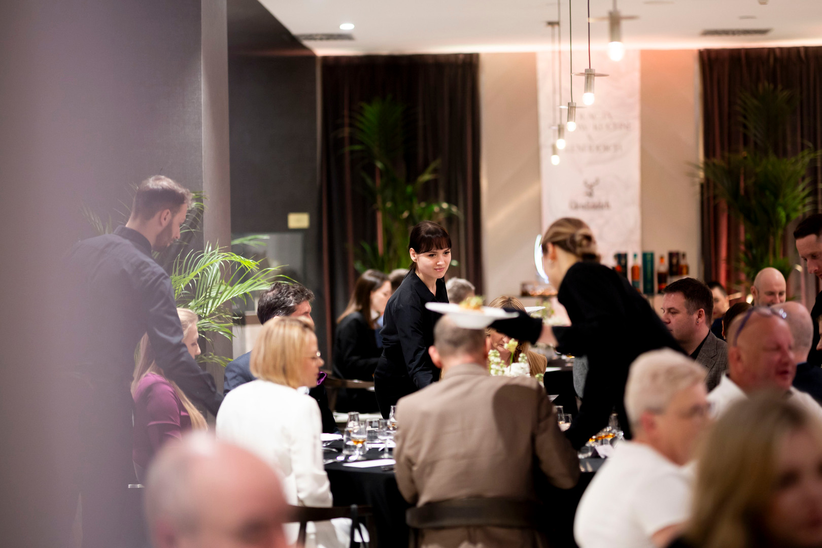 Ruchliwa hotelowa restauracja, personel w czarnych uniformach podaje dania gościom przy ciepłym świetle.