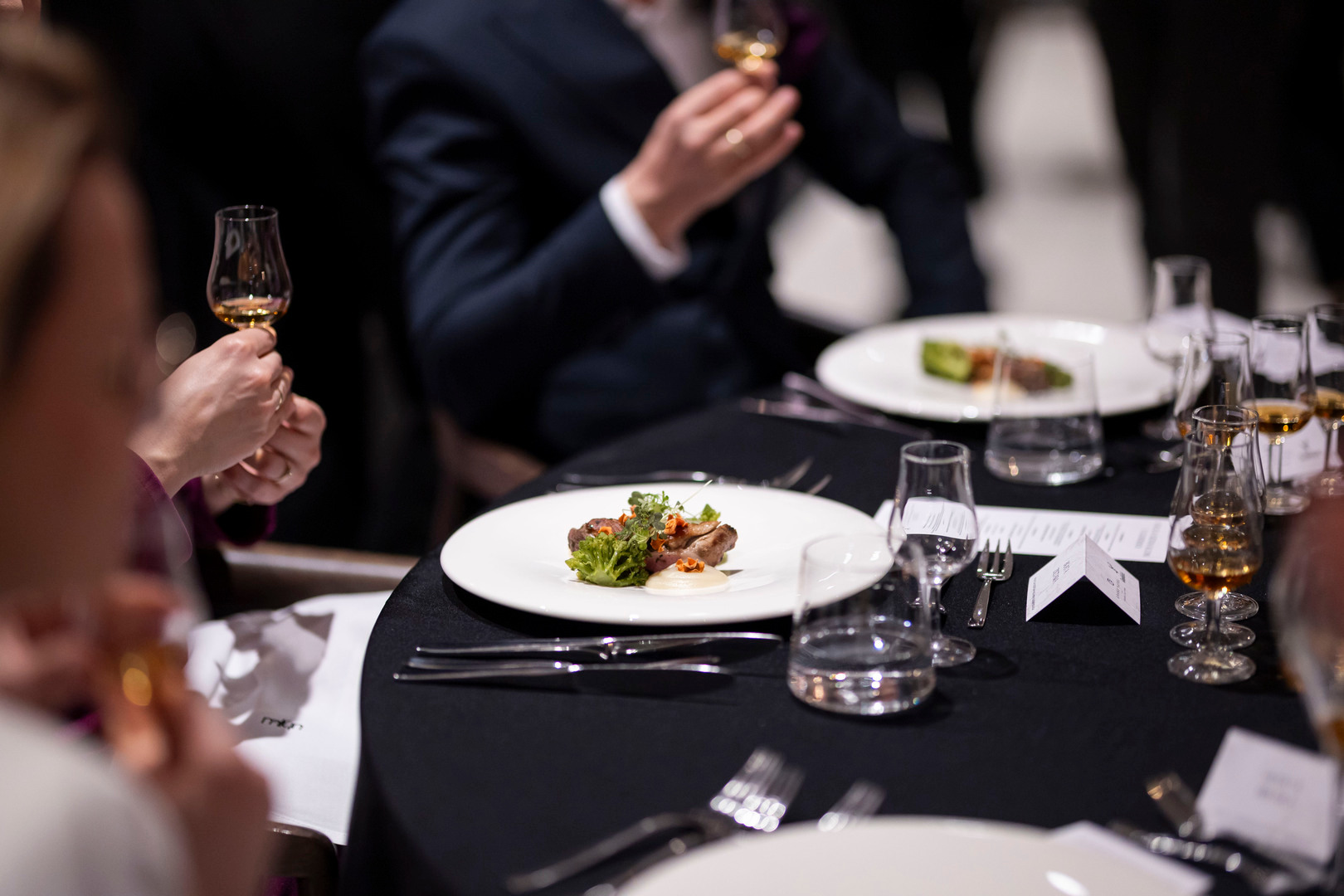 Elegancka kolacja degustacyjna z talerzem przystawki; goście degustują bursztynowe trunki w kieliszkach.