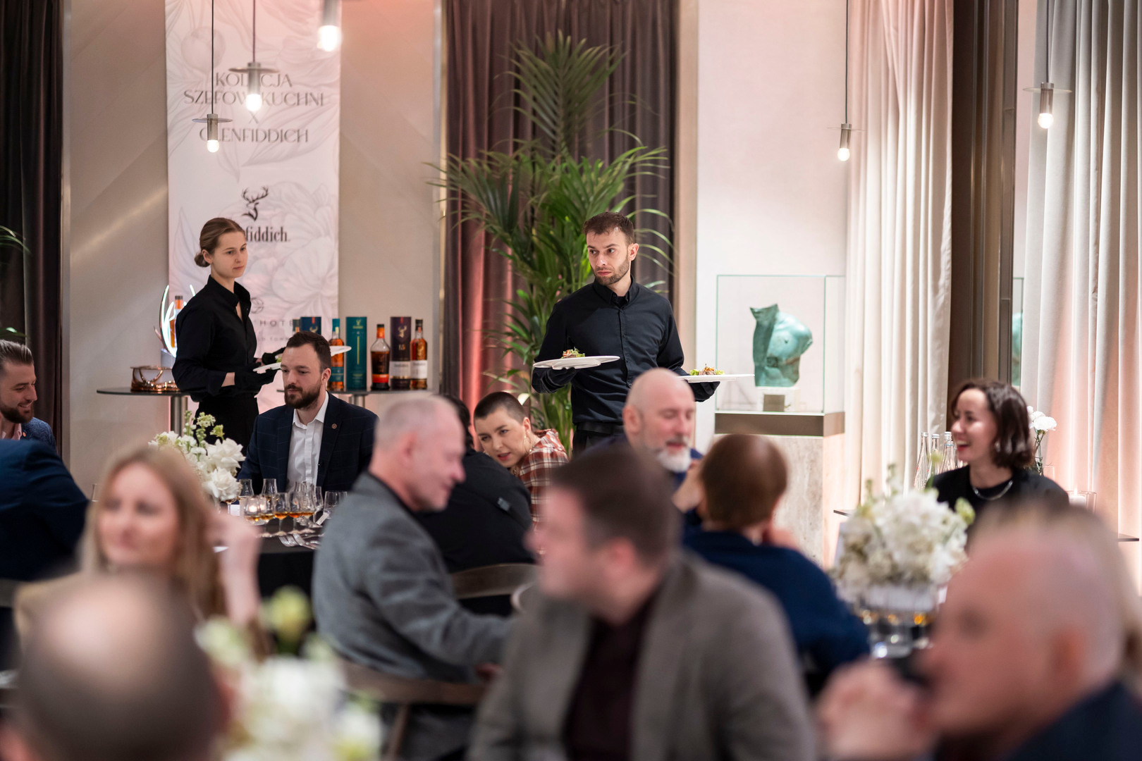 Elegancka sala restauracyjna hotelu, kelnerzy niosą dania, goście przy świecach i kwiatowych dekoracjach.