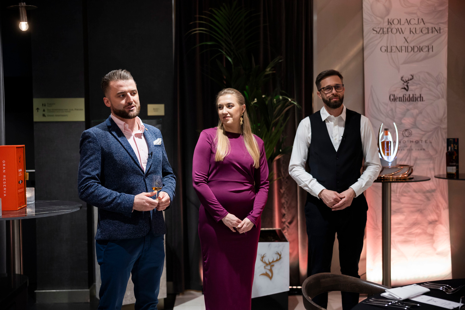 Trójka gospodarzy na eleganckiej degustacji w hotelu: mężczyzna z kieliszkiem whisky, kobieta w fioletowej sukni, obsługa przy stoisku Glenfiddich.