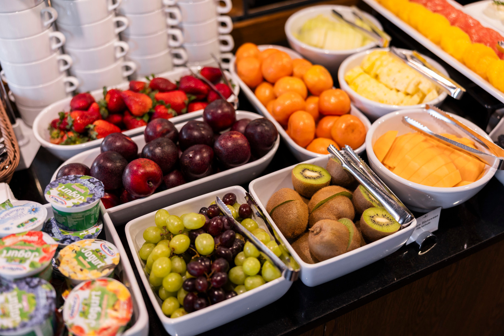 Bufet śniadaniowy hotelu z miskami świeżych owoców — winogrona, kiwi, śliwki, truskawki, mandarynki, mango i jogurty.