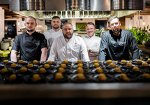 Five uniformed chefs behind a hot-kitchen pass, stainless hood above, rows of plated hors d'oeuvres in foreground.