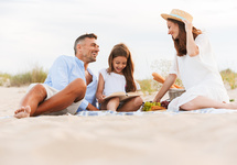 Familienpicknick am Sandstrand mit Decke, Korb, Baguettes und Obst; Eltern lächeln, Kind liest