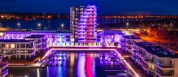 Nowoczesny hotel nad wodą z podświetlaną wieżą, balkonami i przystanią; fioletowo-różowe światła odbijają się w wodzie.