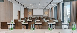 Modernes Hotel-Konferenzzimmer im Schulungsstil mit Leinwand/Projektor, Reihen von Tischen und Stühlen, Wasserflaschen, Notizblöcken und großen Fenstern