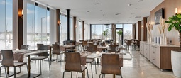 Jasna, nowoczesna restauracja hotelowa z wysokimi oknami, drewnianymi krzesłami, nakrytymi stolikami, ciepłym oświetleniem i zielenią.