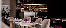 Elegancka restauracja hotelowa z miękkimi fotelami, nakrytymi stołami, fioletowymi kwiatami i podświetlonym barem w tle