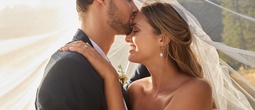 Bride and groom share a tender forehead kiss beneath a flowing veil at a sunlit outdoor wedding.