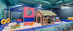 Indoor kids' play area with white ball pit, colorful playhouse, ocean-themed padded panels and climbing frame