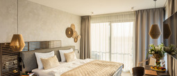 Modern hotel room with king bed, beige quilt, hanging lamps, wooden headboard, sunlit balcony and desk with fruit