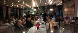 Lang restaurantbord på hotellet med hvite duker, rosa blomster og gjester som spiser under varmt taklys.