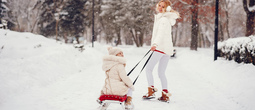 Woman pulls child on a red sled along a snowy park path, both in cozy winter coats, boots and earmuffs as snow falls.