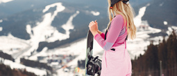 Kobieta w różowym stroju i kasku trzyma deskę snowboardową, podziwiając zaśnieżone stoki i ośrodek