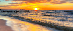 Sunset over a sandy beach with gentle waves and wooden breakwaters, golden sky reflecting on wet sand