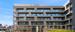 Modern hotel with perforated façade, glass‑railed balconies, ground‑floor terrace seating and young landscaped lawn.