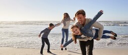 Family laughing on a sandy beachfront with gentle waves, parents lifting children by the shoreline