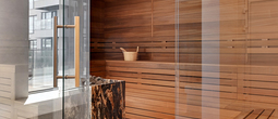 Modern hotel sauna with cedar benches, wooden bucket and ladle, stone heater with marble surround and glass door with brass handle