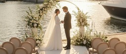 Couple exchanging vows beneath a floral arch on a marina dock at sunset, elegant chairs, candles and yachts in the background