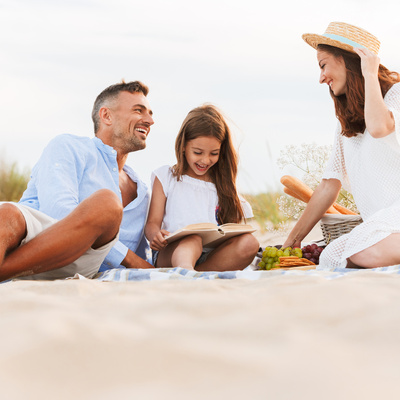 Rodzinny piknik na piaszczystej plaży z kocem, wiklinowym koszem, bagietkami i owocami; rodzice uśmiechają się, dziecko czyta