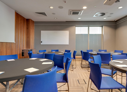 Konferenzraum mit runden Tischen, leuchtend blauen Stühlen, Whiteboard, Projektor, Holzakzentwand und Notizzetteln.