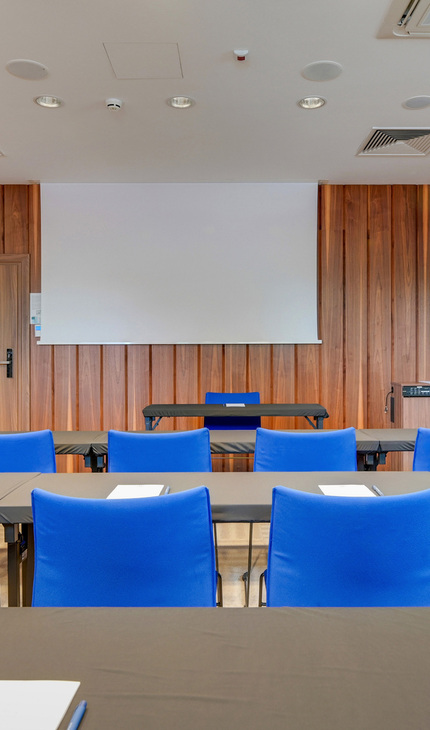 Hotel-Tagungsraum mit blauen Stühlen, langen Tischen, Notizblöcken und Stiften, Projektorleinwand und holzverkleideter Wand.